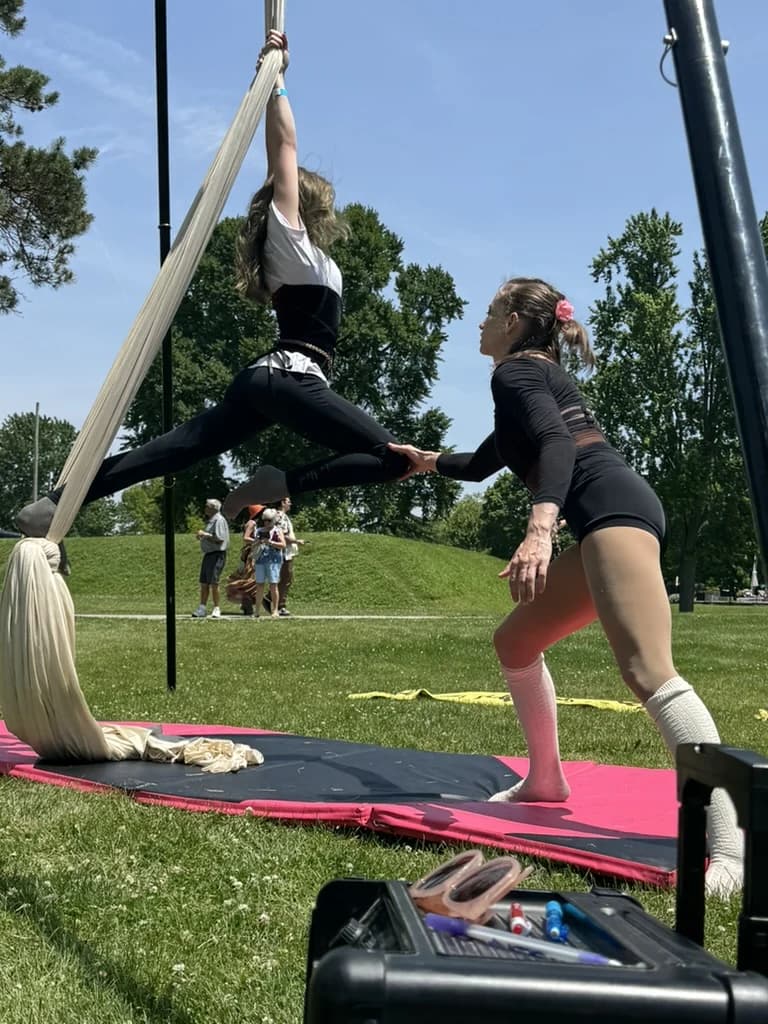 Aerial silks training session — one student suspended in a split, the other spotting from the mat below.